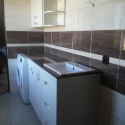 Biała szafka łazienkowa z umywalką i szufladami, obok pralka, na tle brązowo-beżowych płytek i wiszącej szafki z półkami.