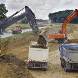 Żółta i pomarańczowa koparka ładują urobek do wywrotek na placu budowy drogi, w tle ruch samochodowy na nowej trasie.