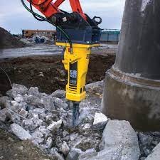 Hydrauliczny młot wyburzeniowy zamontowany na ramieniu koparki, kruszący betonowe elementy na placu budowy, z widocznym fragmentem betonowej rury w tle.