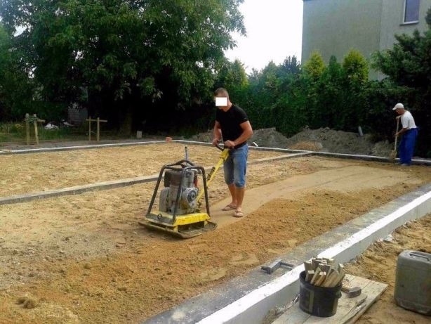 Pracownik zagęszcza piasek zagęszczarką płytową w przygotowanym wykopie pod fundamenty, drugi pracownik wyrównuje teren łopatą w tle.