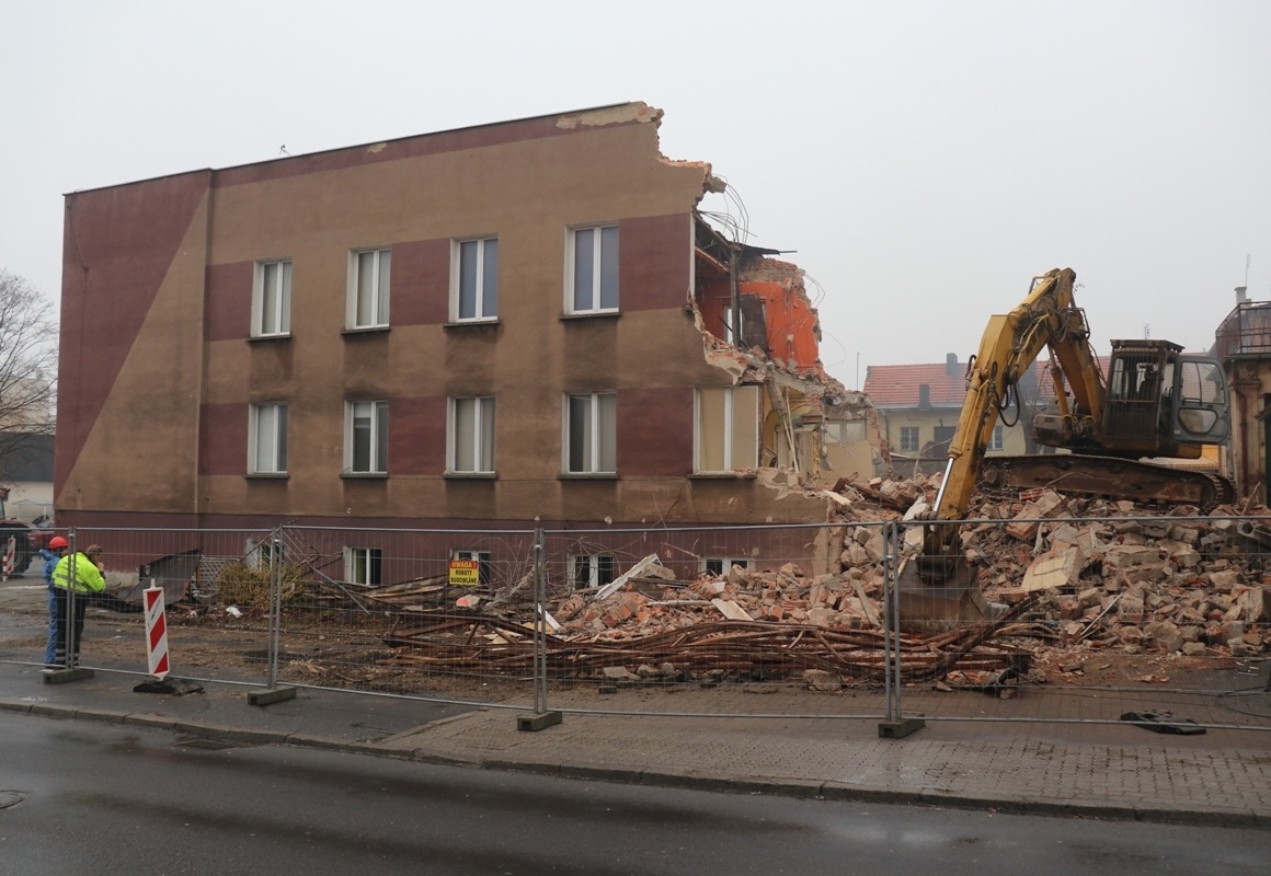 Żółta koparka przy częściowo zburzonym, czteropiętrowym budynku z widocznymi cegłami, otoczonym tymczasowym ogrodzeniem i znakami ostrzegawczymi.