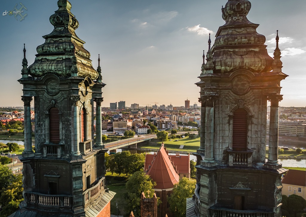 Widok z góry na Bazylikę Archikatedralną św. Apostołów Piotra i Pawła we Wrocławiu, z dwiema wieżami na pierwszym planie i panoramą miasta w tle, uchwycony w ciepłym świetle zachodzącego słońca.