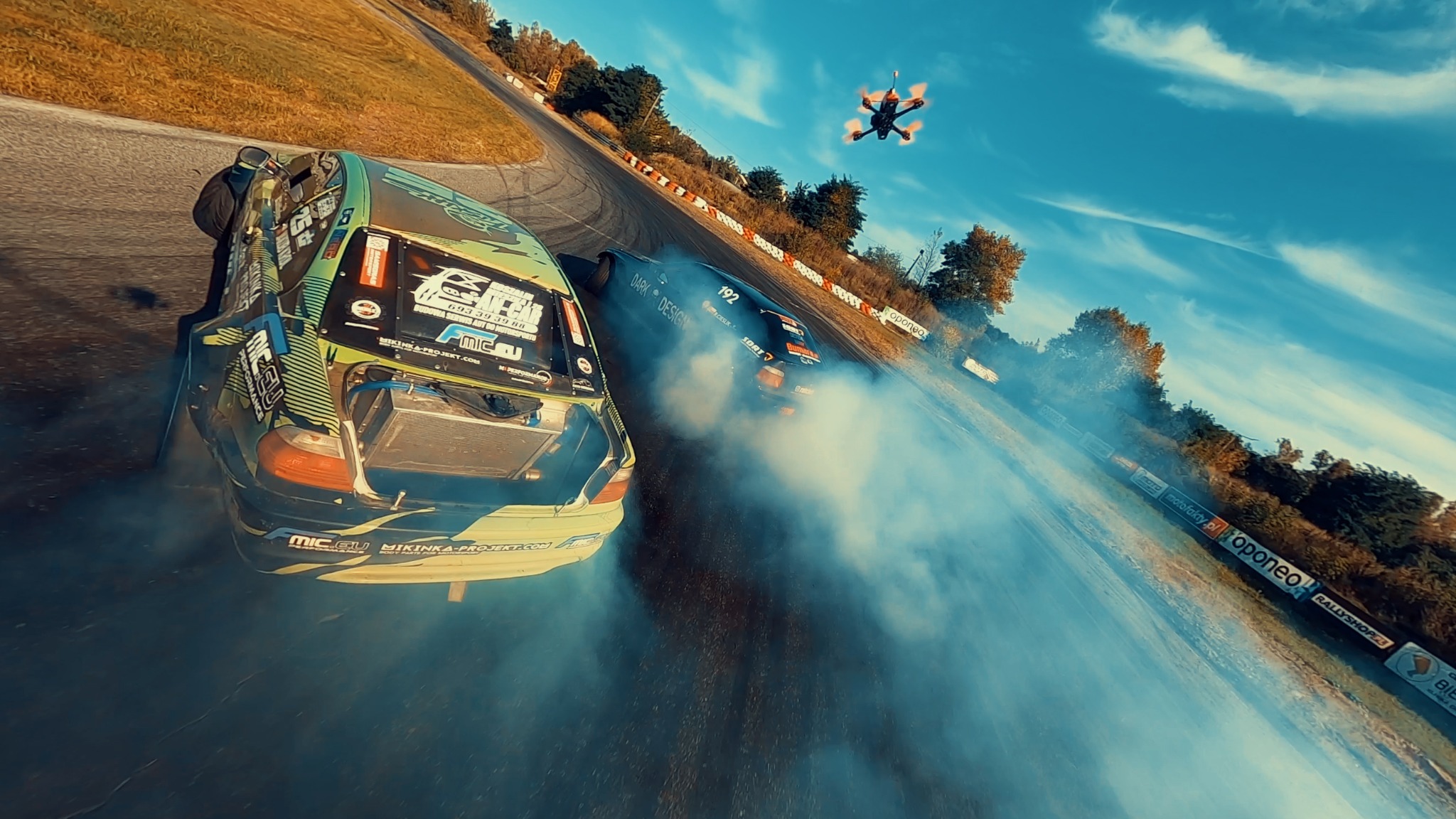 Dwa samochody driftujące na torze wyścigowym, ujęcie z drona FPV, widoczny dym z opon i reklamy na bandach toru.