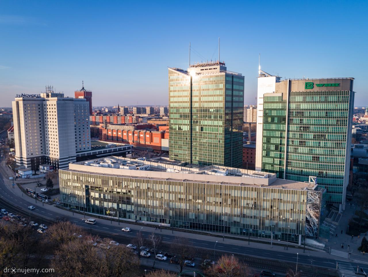 Panorama miasta z wysokimi biurowcami o szklanych fasadach w słoneczny dzień, widok z góry na ruch uliczny i zaparkowane samochody.