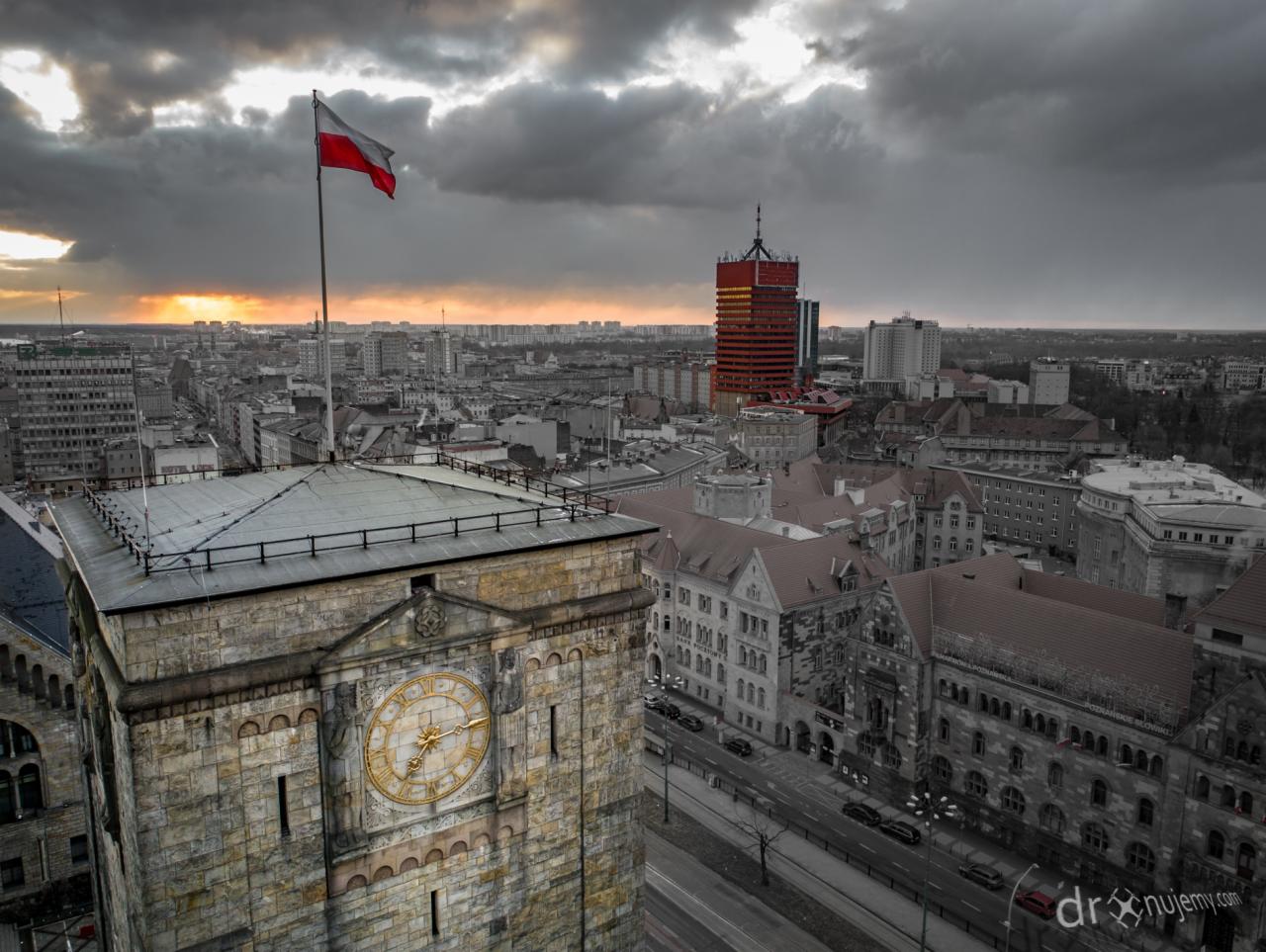 Panorama miasta z wieżą zegarową i powiewającą polską flagą na tle zachodzącego słońca i burzowych chmur.