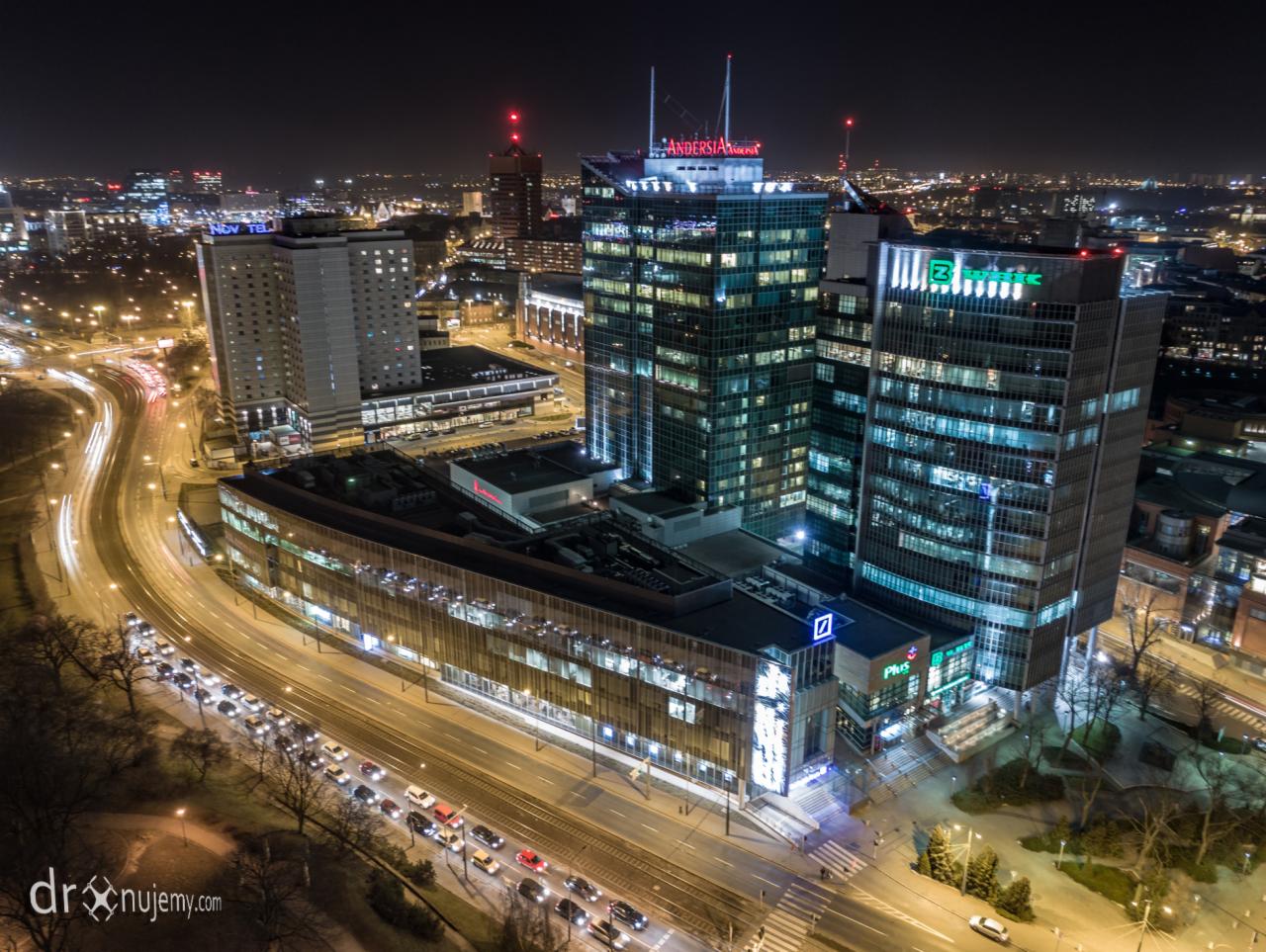 Nocny widok z lotu ptaka na kompleks biurowców z podświetlonymi logo firm, długie ekspozycje świateł samochodów na ulicach.