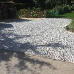 Utwardzony szarym żwirem teren w ogrodzie, obok kostka brukowa i drewniane ogrodzenie, w tle drzewa.