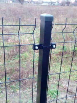 Fragment czarnego ogrodzenia panelowego z metalową siatką i słupkiem zakończonym czarną nakładką, mocowanym na dwie śruby, na tle łąki.