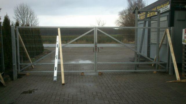 Podwójna, metalowa brama z siatki w trakcie montażu, podparta drewnianymi stemplami i poziomnicą na kostce brukowej, widok z zewnątrz.