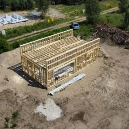 Wysoka perspektywa na drewniany szkielet domu w budowie, z widocznymi fundamentami i przyczepą na piaszczystym terenie.