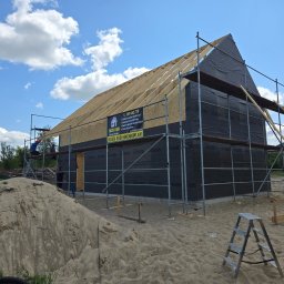 FACH BUD USŁUGI REMONTOWO - BUDOWLANE TOMASZ CHOMICZ - Budowa domu w trakcie: widoczne drewniane krokwie dachu, ściany pokryte membraną, rusztowania i baner reklamowy firmy budowlanej. Na pierwszym planie hałda piasku.