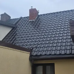 Błyszczący, ciemny dach z dachówki ceramicznej na domu z beżową elewacją. Widoczny komin z cegły i orynnowanie w kolorze dachu. Pochmurne niebo w tle.