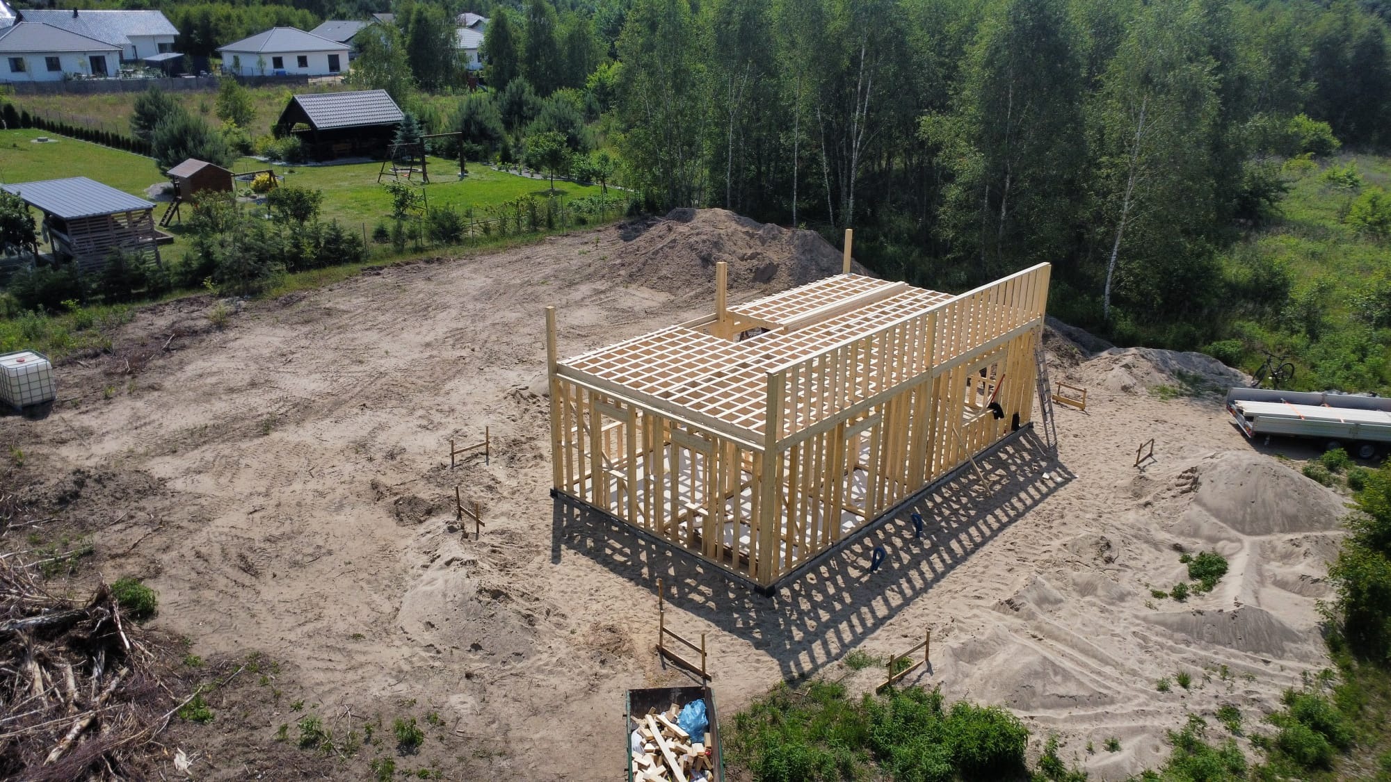 Drewniana konstrukcja szkieletowa domu w trakcie budowy, widok z góry, teren piaszczysty, w tle las i zabudowania.