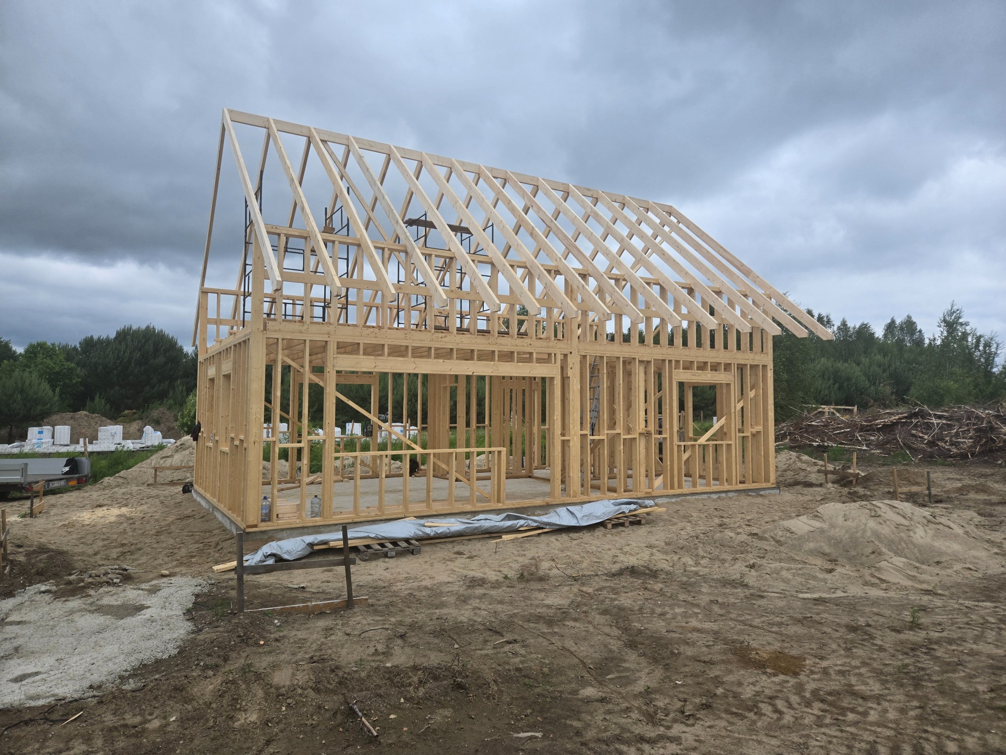Surowy szkielet domu z drewnianą konstrukcją dachu, widok z zewnątrz na tle pochmurnego nieba. Budowa w toku, widoczne elementy konstrukcyjne i teren budowy.