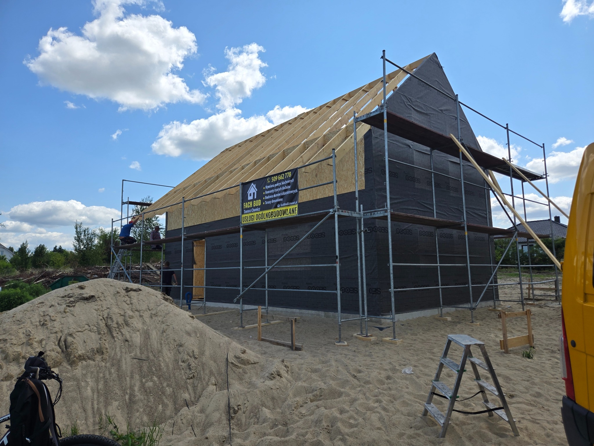 Budowa domu w trakcie: widoczne drewniane krokwie dachu, ściany pokryte membraną, rusztowania i baner reklamowy firmy budowlanej. Na pierwszym planie hałda piasku.