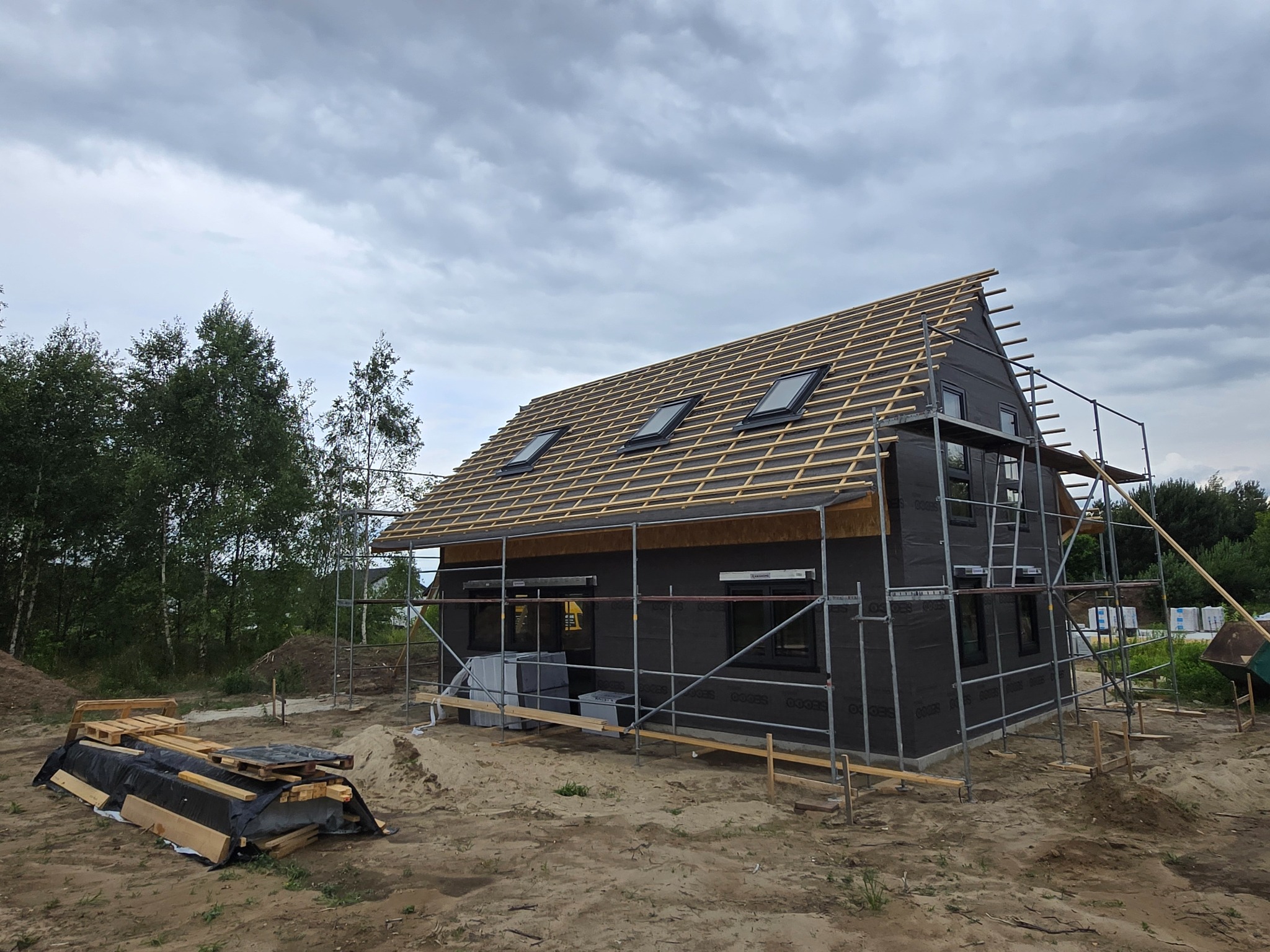 Budowa domu jednorodzinnego w trakcie krycia dachu. Widoczne rusztowania, okna dachowe i konstrukcja więźby. Dom z czarną membraną na ścianach.