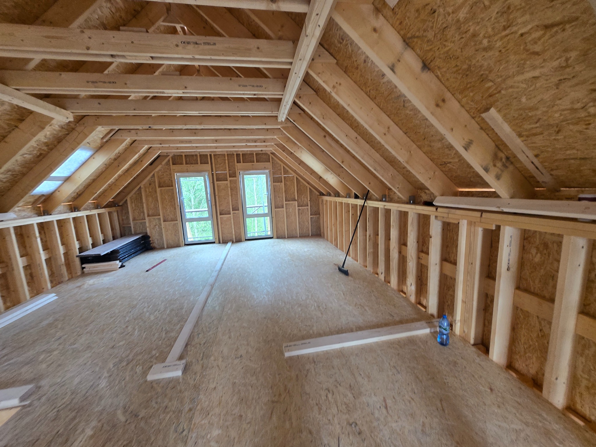 Poddasze z drewnianą konstrukcją dachu w trakcie budowy. Widoczne okna, elementy drewniane i narzędzia. Charakterystyczny wzór płyty OSB na ścianach i podłodze.