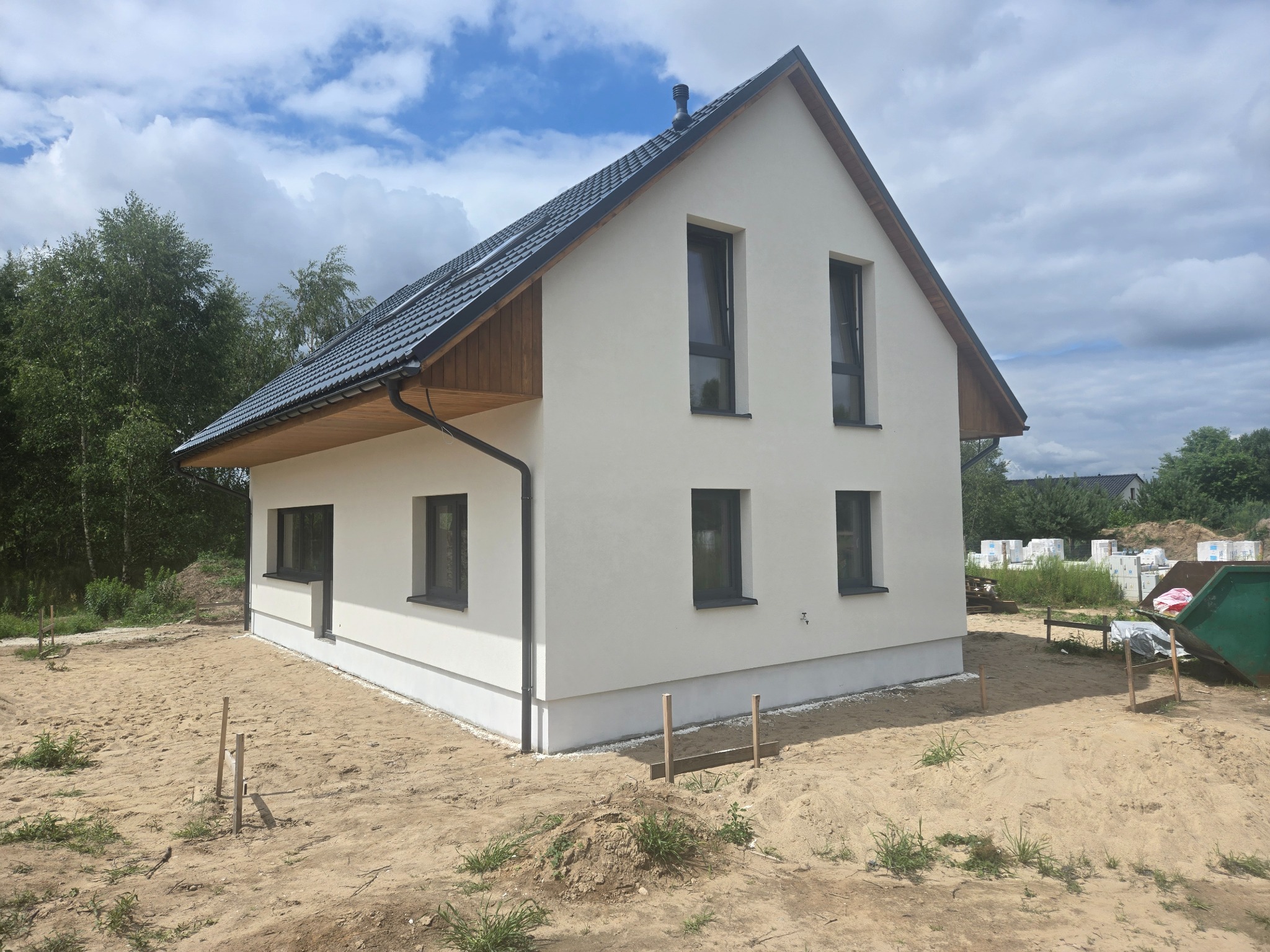 Nowoczesny dom jednorodzinny z grafitowym dachem i jasną elewacją, w otoczeniu piaszczystego terenu i zieleni. Widoczne okna w ciemnych ramach i elementy wykończenia dachu.