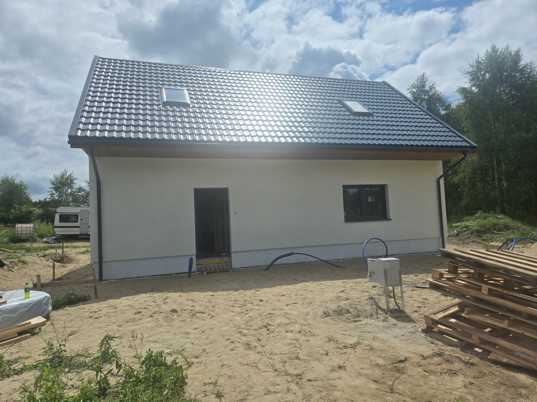 Dom jednorodzinny z nowym, błyszczącym, szarym dachem z dachówką metalową, oknami dachowymi i białymi ścianami. W tle drzewa i błękitne niebo z chmurami.