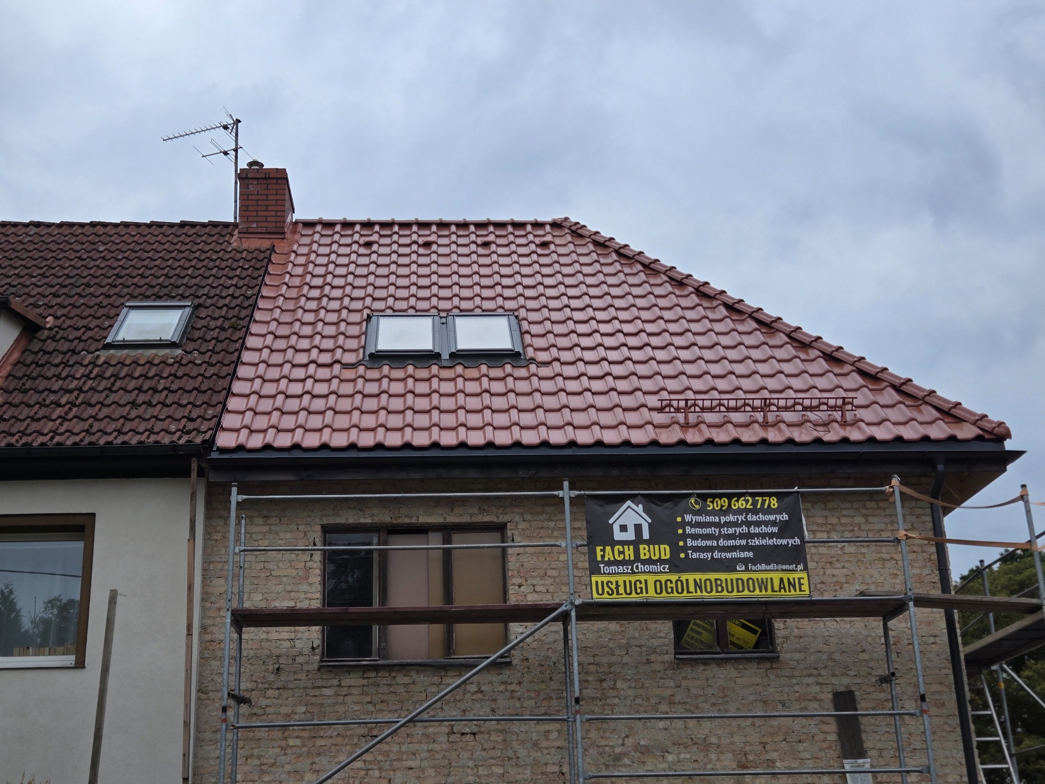 Częściowy widok domu z nową, czerwoną dachówką i oknami dachowymi. Na pierwszym planie rusztowanie i baner reklamowy firmy budowlanej.