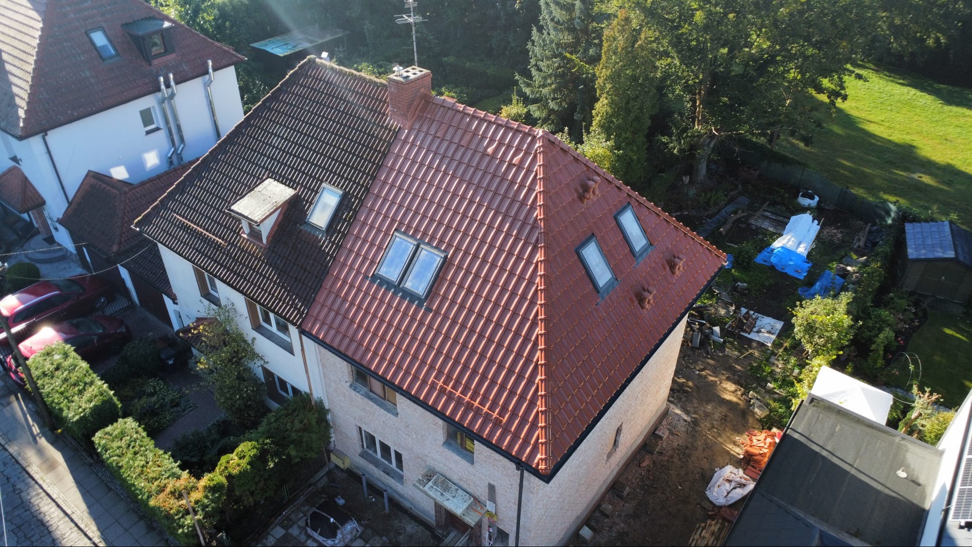 Dom z częściowo wymienionym dachem z czerwonej dachówki, widok z góry. Stara, ciemna dachówka kontrastuje z nową, jasną. Okna dachowe w obu częściach dachu.