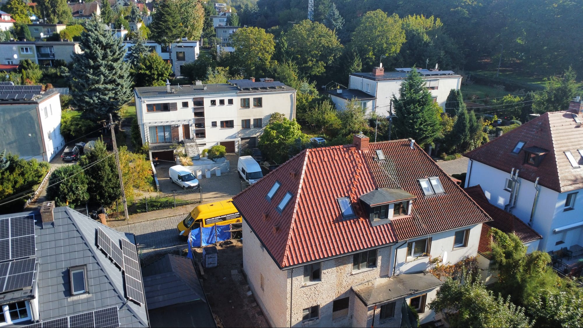 Czerwony dach z oknami dachowymi po renowacji, widok z góry na tle zieleni i zabudowy miejskiej. Żółty bus serwisowy pod plandeką obok budynku.