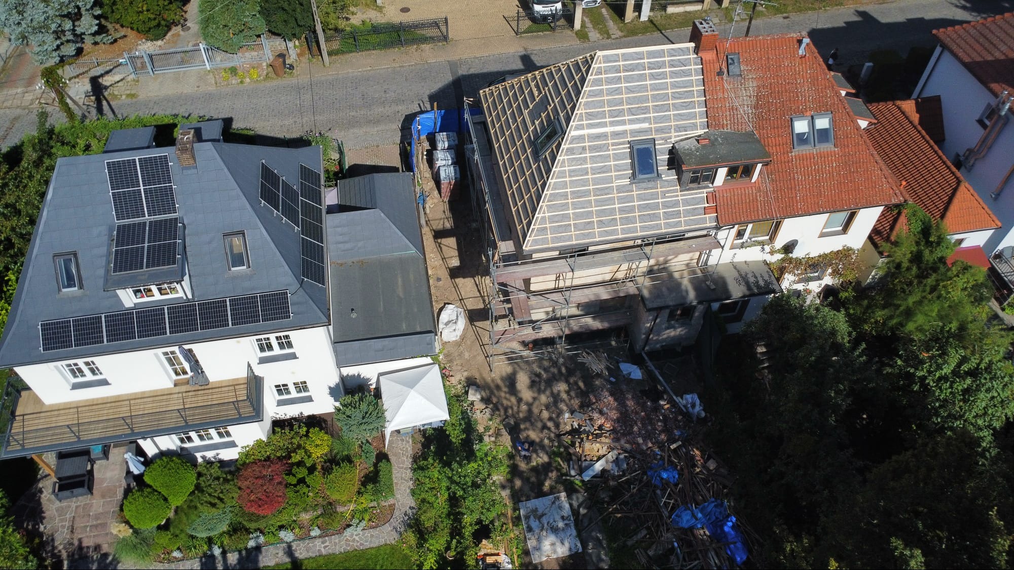 Widok z góry na dom w trakcie wymiany dachu. Częściowo zdjęte stare dachówki, widoczna konstrukcja więźby i rusztowania. Obok dom z panelami słonecznymi na dachu.