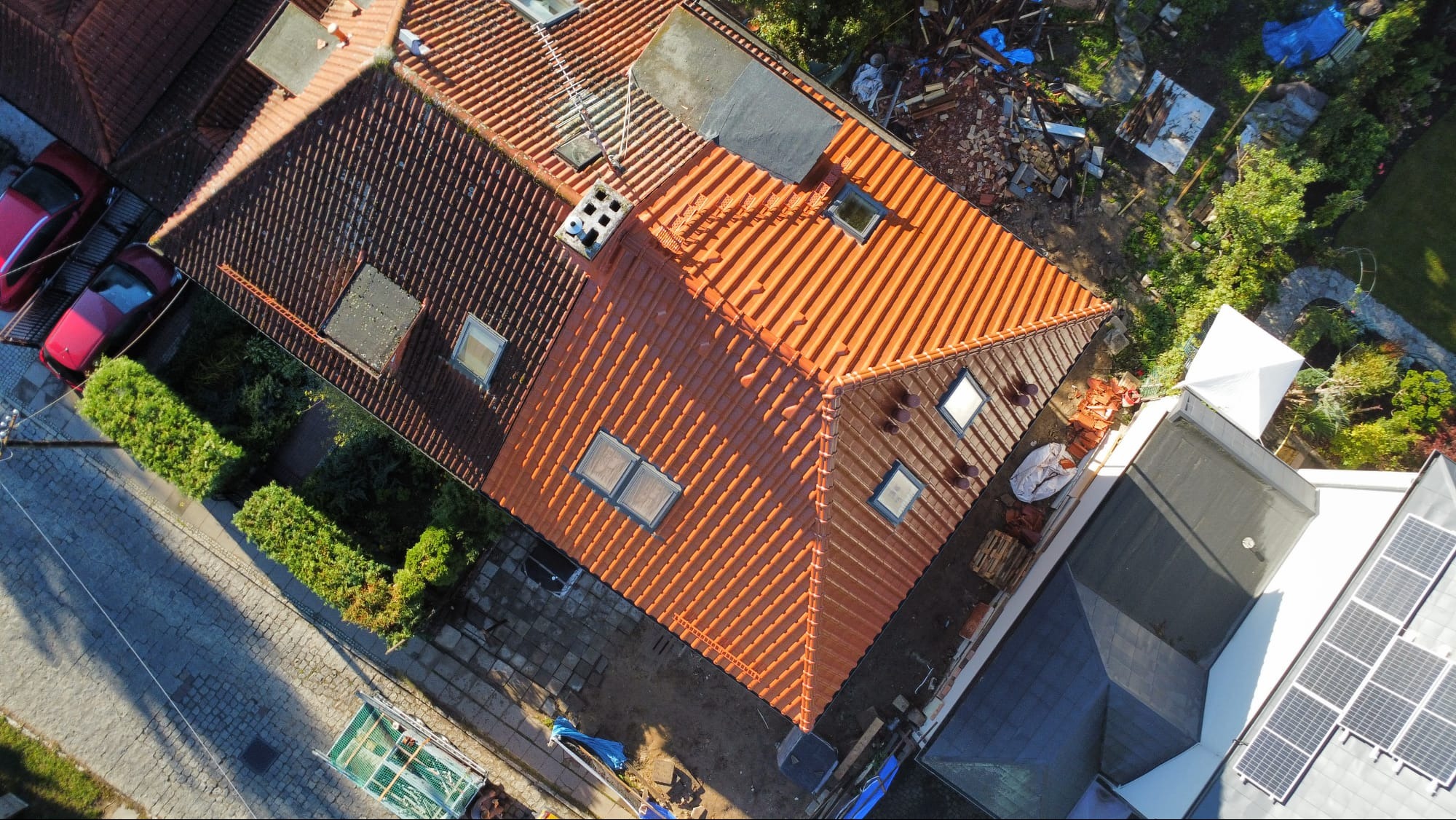 Widok z góry na dom z częściowo wymienionym dachem z pomarańczowej dachówki, obok starej, ciemnej dachówki. Widoczne okna dachowe, panele słoneczne i dwa czerwone samochody.