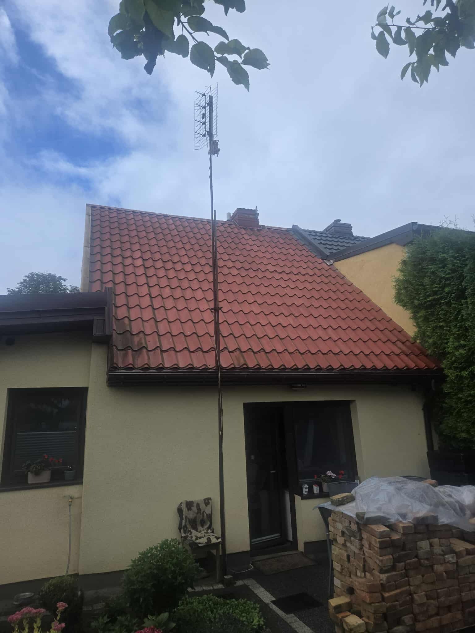 Dom jednorodzinny z nowym, czerwonym dachem z dachówki ceramicznej. Na pierwszym planie sterta cegieł przy wejściu. Antena na dachu. Widok z poziomu gruntu.