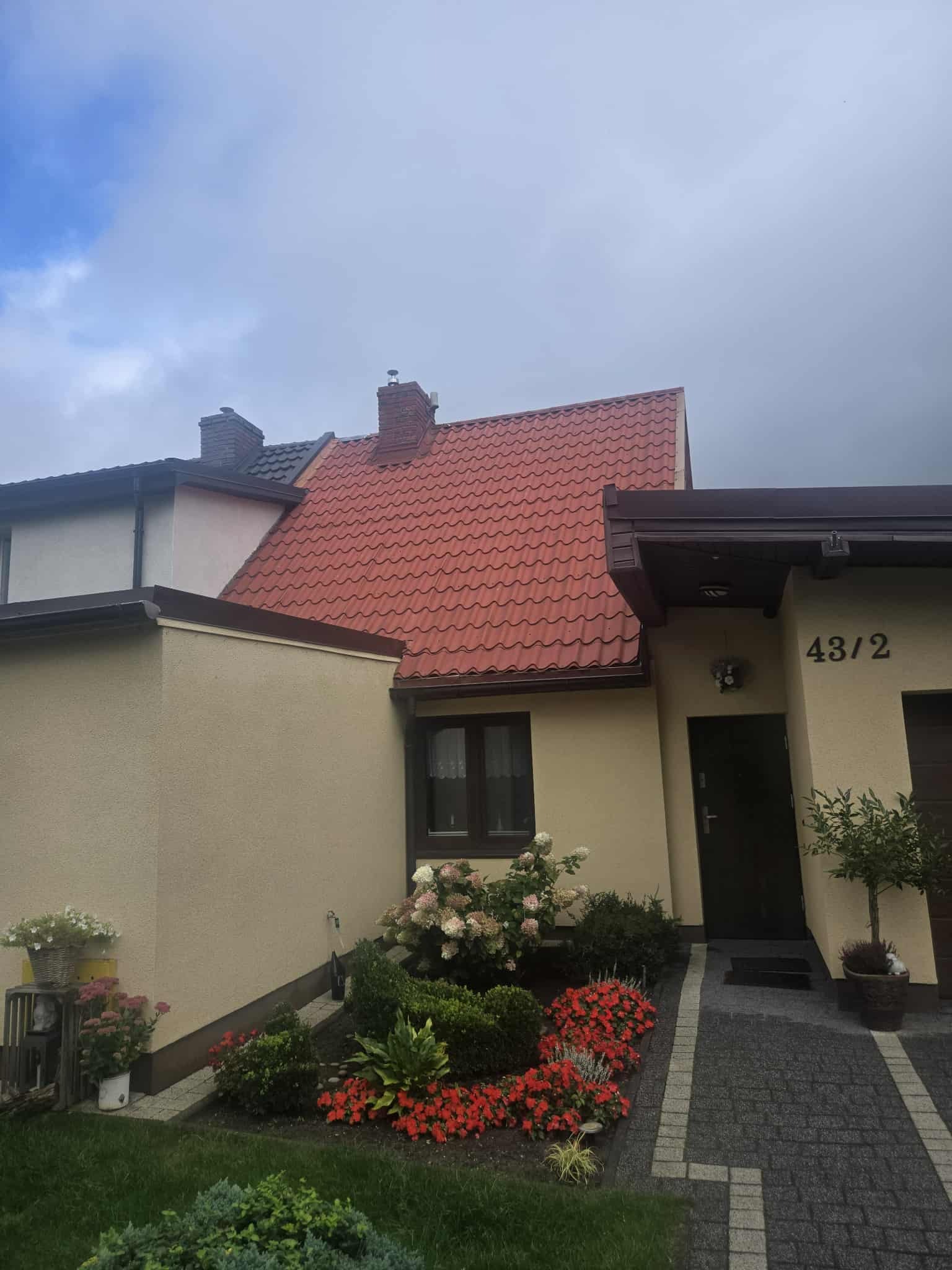 Dom jednorodzinny z nowym, czerwonym dachem z dachówki ceramicznej. Widoczne detale wykończenia, zadbany ogród z kwiatami i krzewami. Numer domu 43/2.