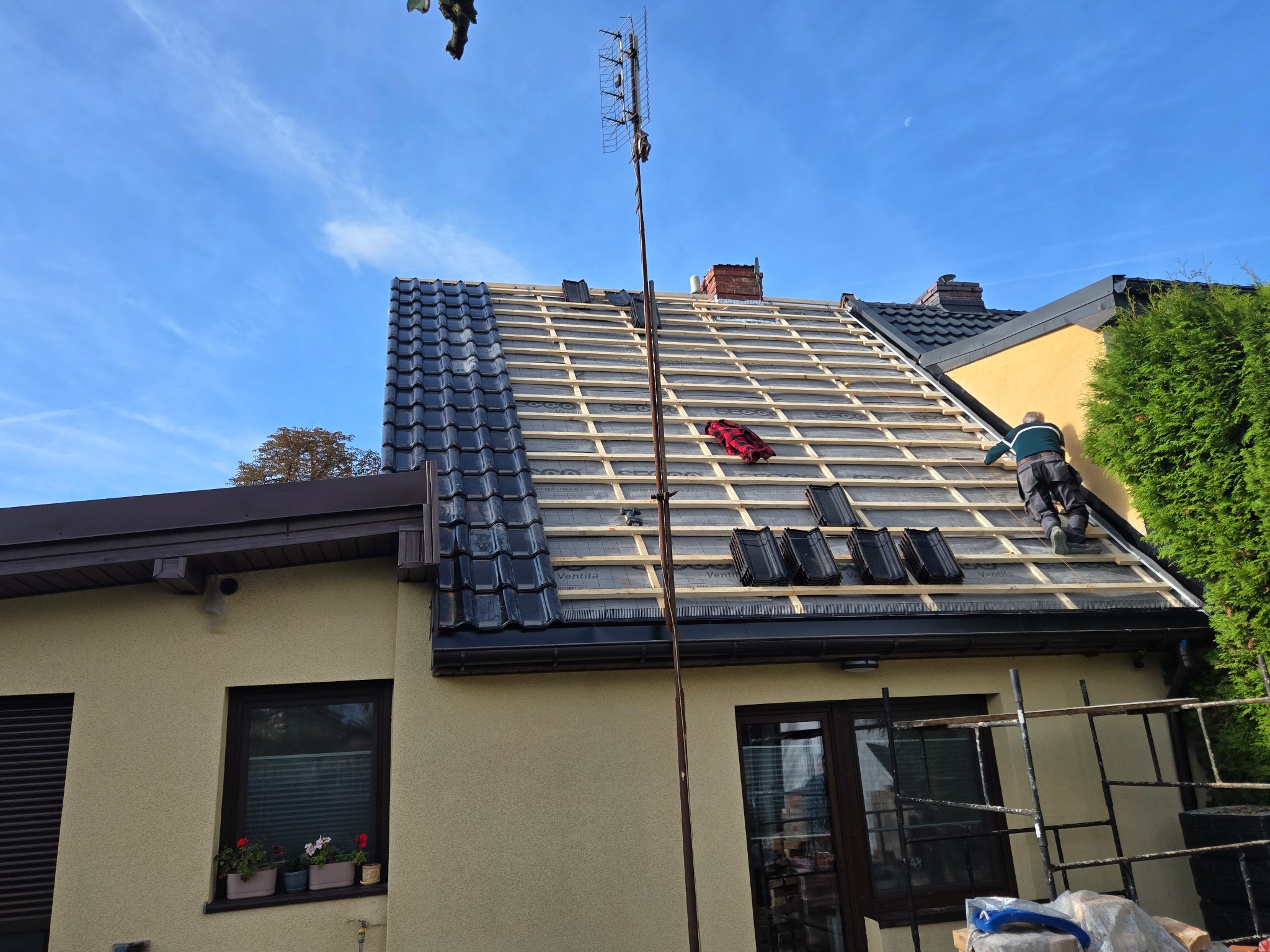 Układanie czarnej dachówki na drewnianej konstrukcji dachu domu jednorodzinnego. Widoczny dekarz w trakcie pracy, antena i fragment elewacji budynku.