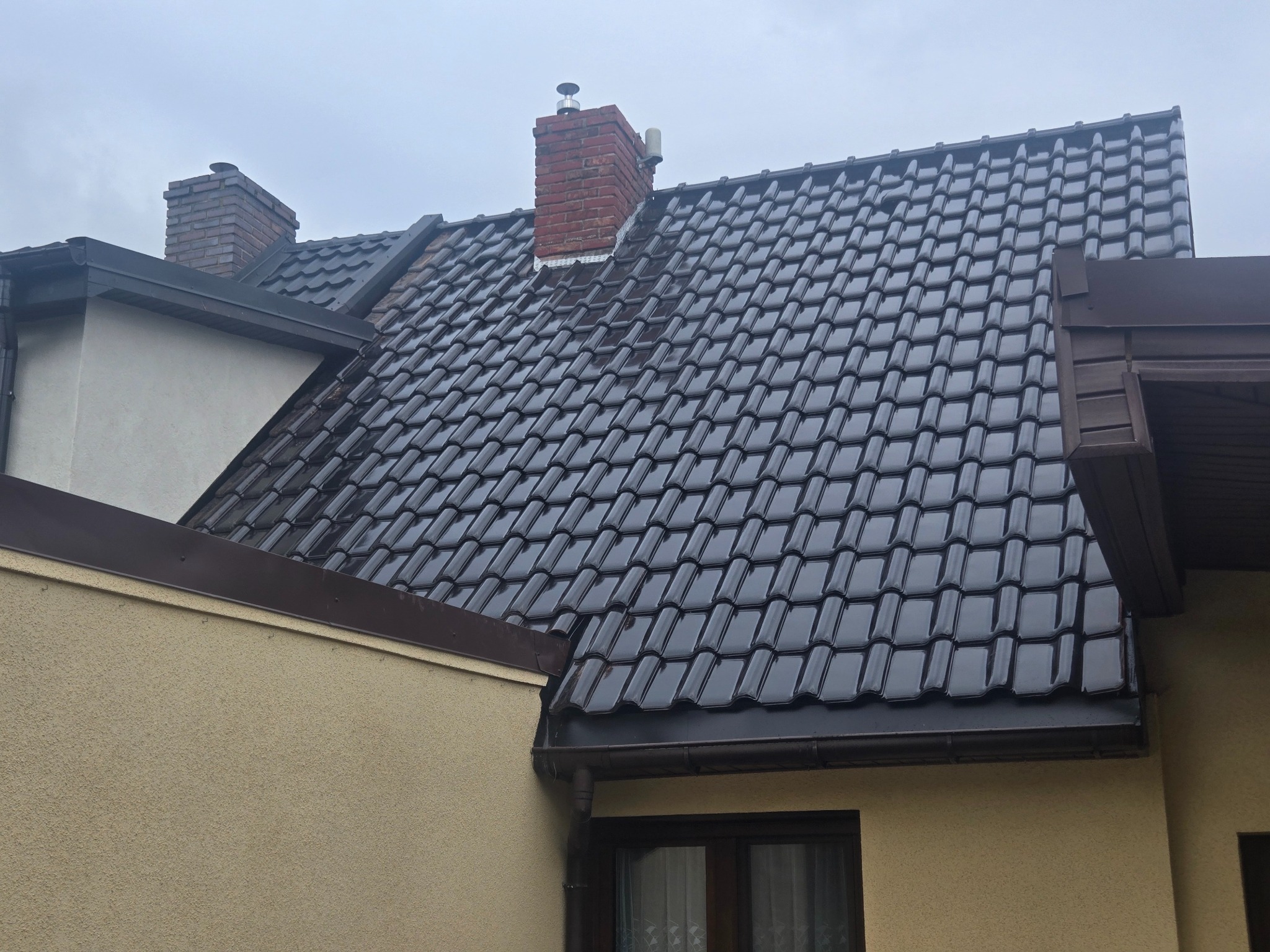 Błyszczący, ciemny dach z dachówki ceramicznej na domu z beżową elewacją. Widoczny komin z cegły i orynnowanie w kolorze dachu. Pochmurne niebo w tle.