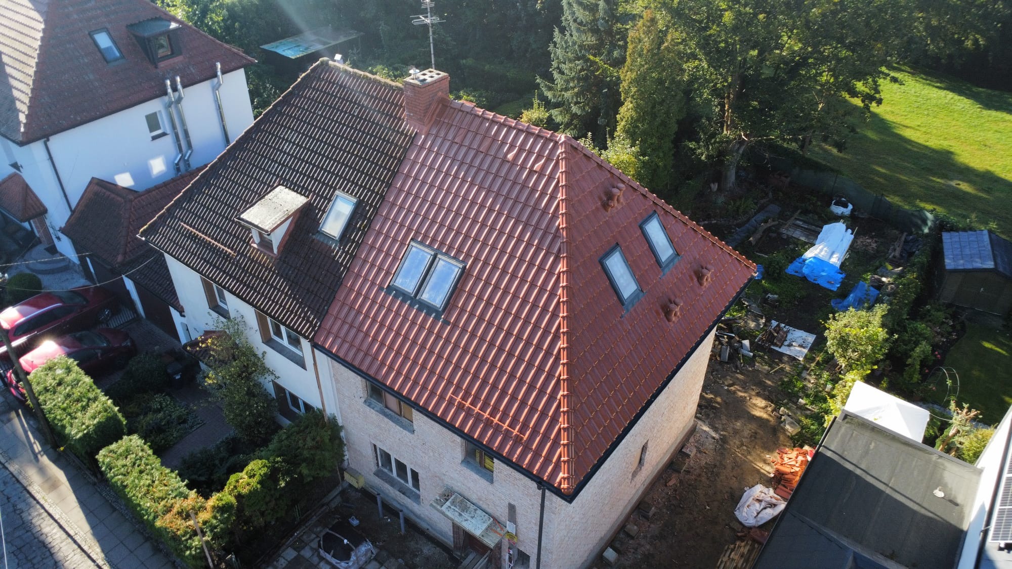 Dom z częściowo wymienionym dachem z czerwonej dachówki, widok z góry. Stara, ciemna dachówka kontrastuje z nową, jasną. Okna dachowe w nowej części dachu.
