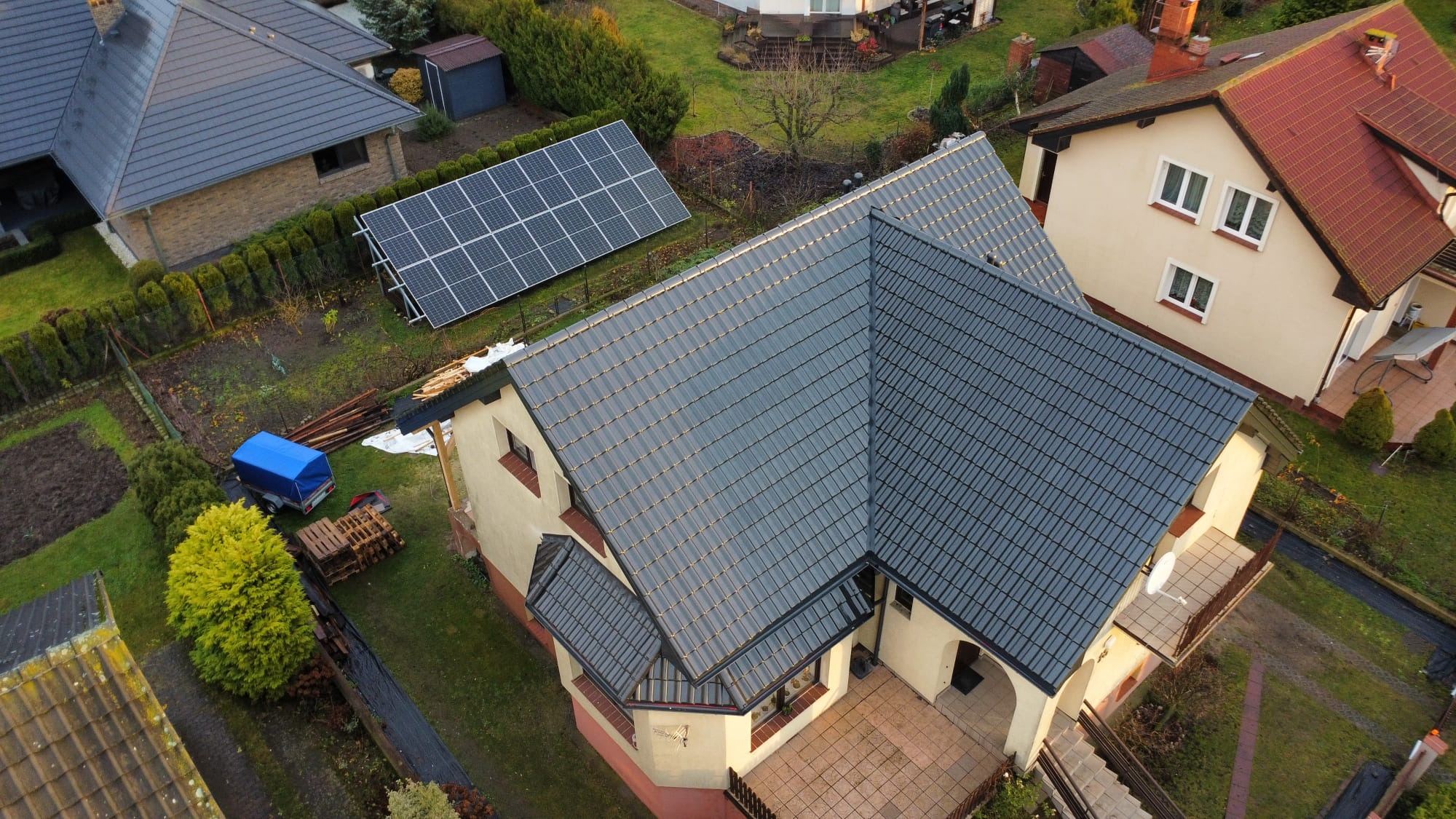 Dom z nowym, grafitowym dachem z dachówki ceramicznej, widok z góry. Obok panele słoneczne i przyczepka z plandeką. Estetyczne wykończenie.