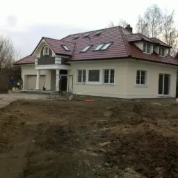 Niedokończony dom jednorodzinny z czerwoną dachówką, kremową elewacją, białymi oknami i kolumnami przy wejściu, widoczny fragment murowanej konstrukcji i rusztowanie, teren wokół domu...