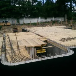 Zbrojony fundament budynku w trakcie budowy, widoczne drewniane szalunki, stalowe pręty zbrojeniowe i wykop ziemny w tle z zielenią.