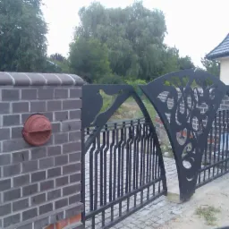 Otwarta, metalowa brama wjazdowa z ozdobnymi elementami, osadzona na murowanych słupkach z cegły klinkierowej, z zamontowaną skrzynką na listy.