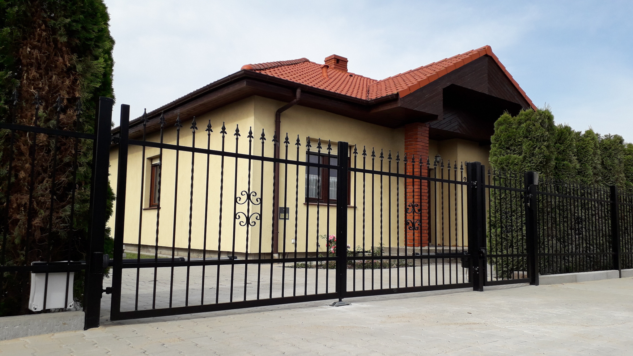 Czarna, metalowa brama wjazdowa z dekoracyjnymi elementami na tle domu z czerwoną dachówką i zadbanym żywopłotem.