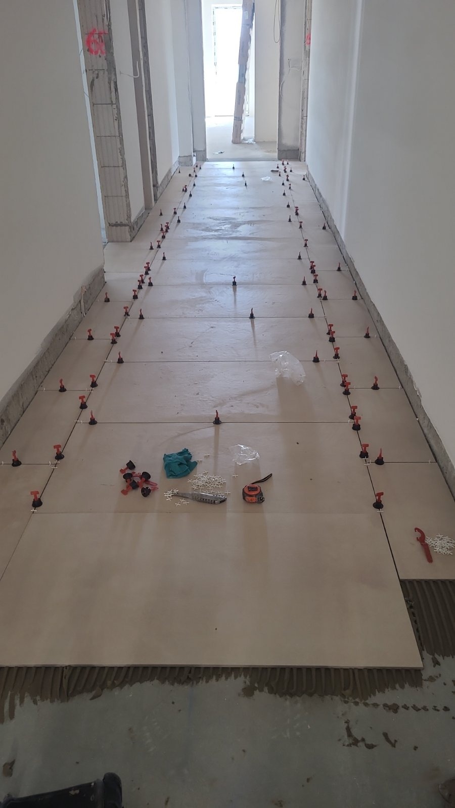 Korytarz w trakcie prac: ułożone beżowe płytki z systemem poziomowania, widoczne narzędzia i klej. Oświetlone wnętrze, perspektywa w głąb pomieszczenia.