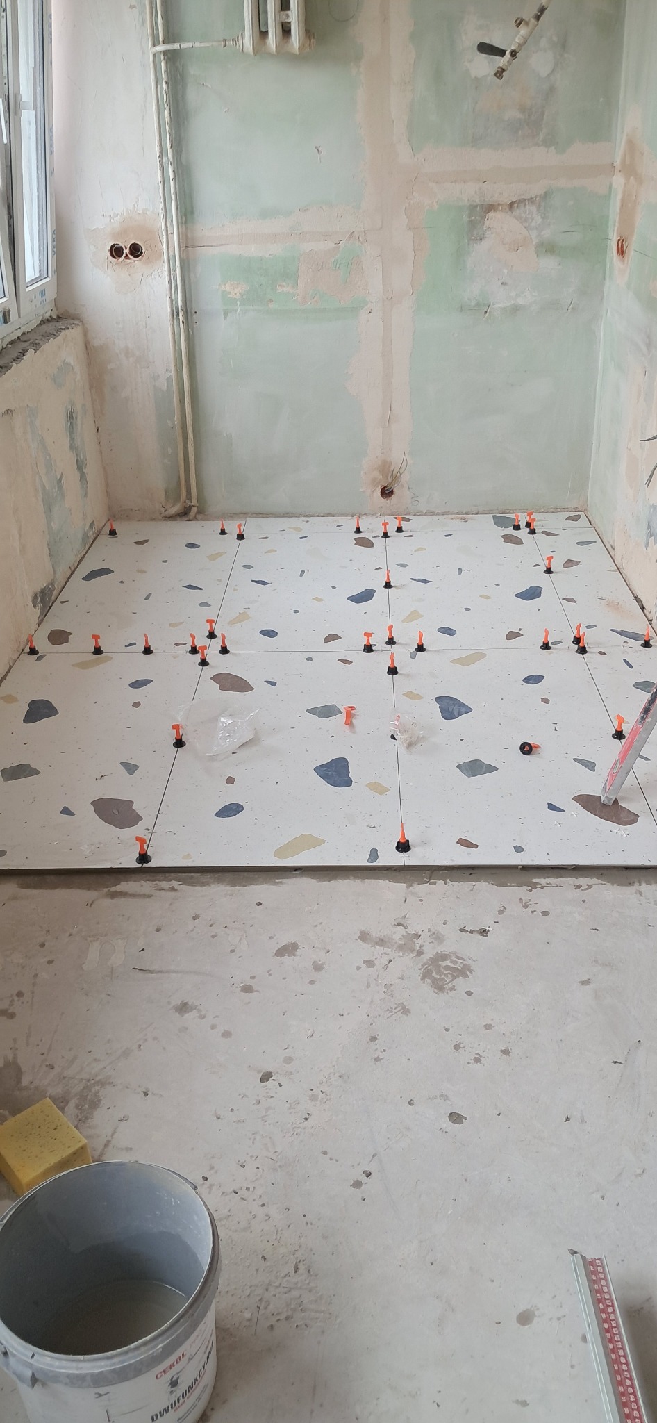 Wnętrze w trakcie prac: ułożone płytki podłogowe w mozaikę, z systemem poziomowania. Ściany surowe, przygotowane do dalszych prac. Widoczny kubeł i gąbka.