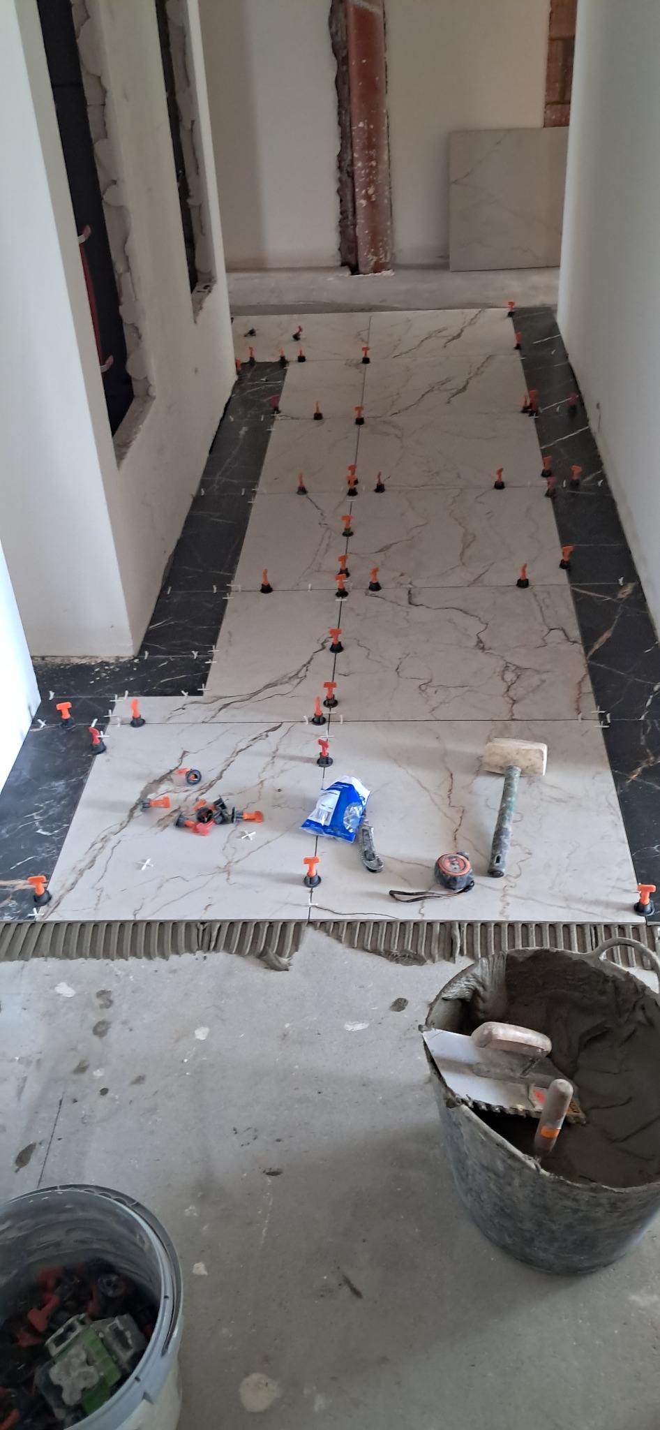 Układanie marmurowych płytek podłogowych z użyciem systemu poziomowania. Widoczne narzędzia: młotek, miarka, paczka z klinami. Fragment korytarza w trakcie prac.