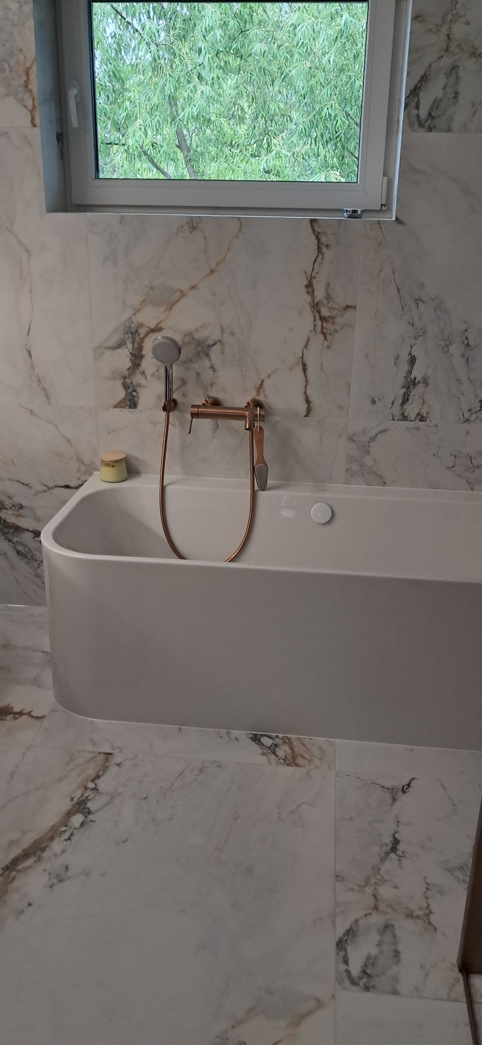 Nowoczesna, biała wanna z armaturą w odcieniach miedzi na tle marmurowych płytek w łazience w Kleosinie. Widok na okno z zielenią.