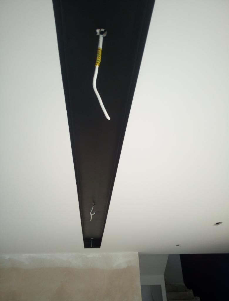 Biały sufit z czarną, wpuszczoną listwą oświetleniową w kształcie litery V, widoczne przewody elektryczne, ściana w trakcie wykończenia na dole kadru, schody w tle.