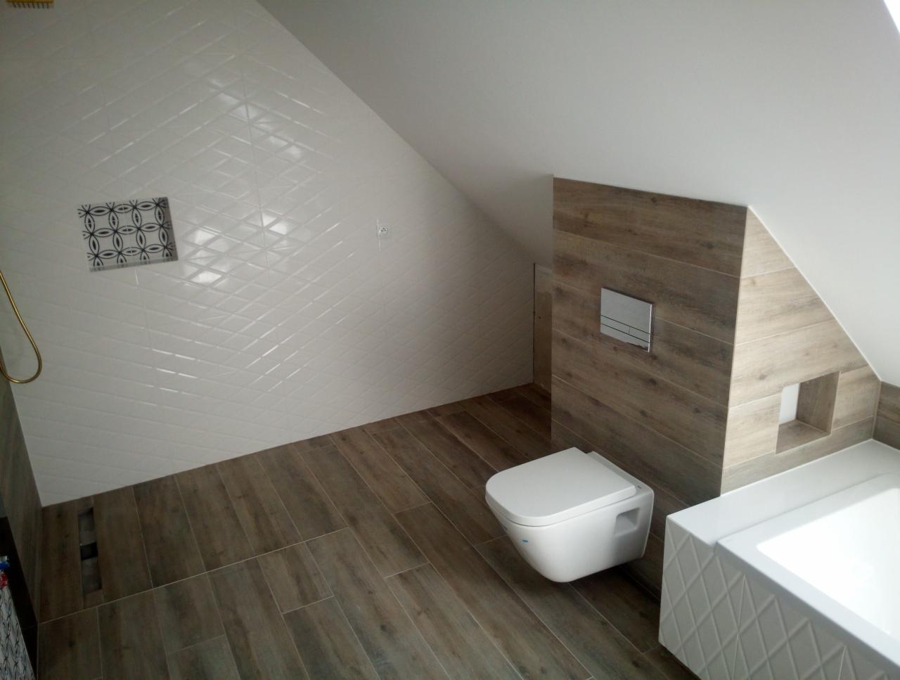 Nowoczesna łazienka na poddaszu ze skośnym sufitem, białe płytki w romby na ścianach, drewnopodobne płytki na podłodze i zabudowie WC, wisząca toaleta i wanna z dekoracyjną obudową.