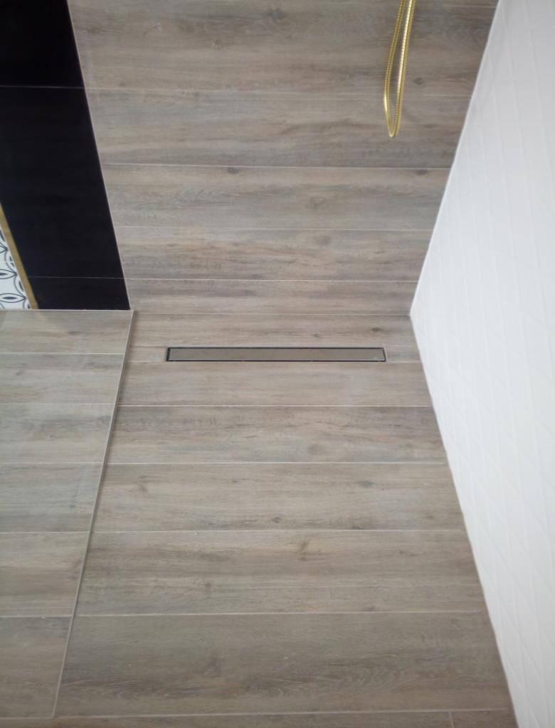 Wykończona podłoga prysznicowa z płytek imitujących drewno, z odpływem liniowym i białymi płytkami na ścianie, widok z góry.