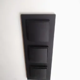 Czarna, pionowa obudowa z trzema prostokątnymi klapkami na białej ścianie, prawdopodobnie element instalacji elektrycznej lub teletechnicznej.
