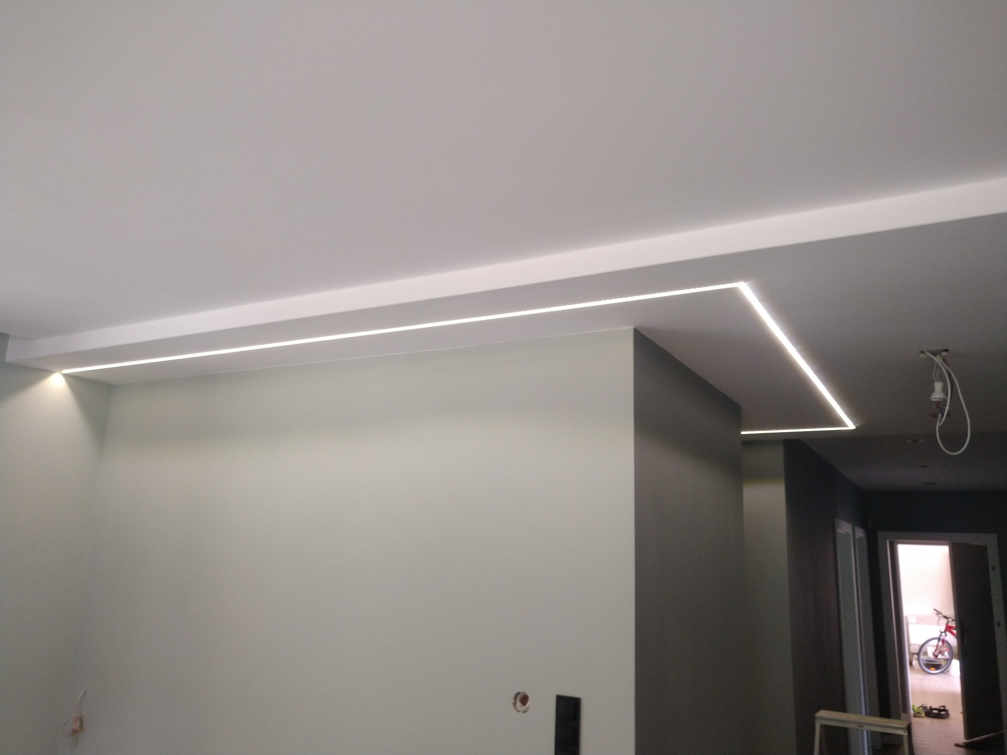 Nowoczesne oświetlenie sufitowe LED w kształcie litery L, w minimalistycznym wnętrzu z szarą ścianą i widocznymi gniazdkami elektrycznymi.