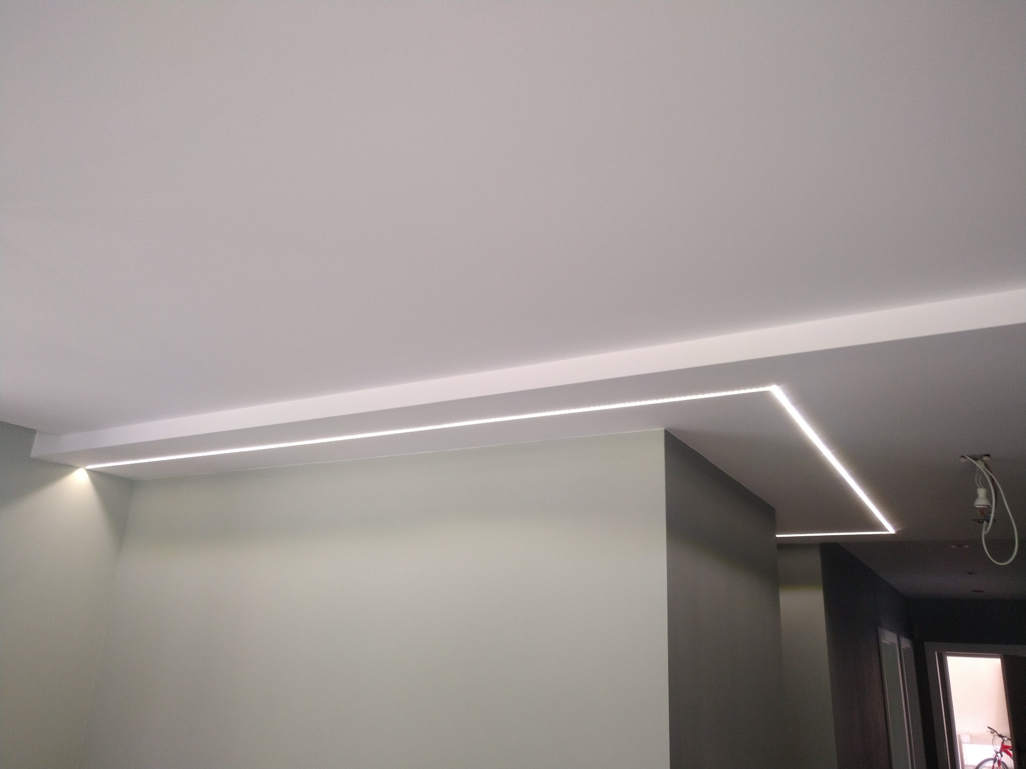 Nowoczesne oświetlenie sufitowe LED w kształcie litery L, z delikatnym światłem podkreślającym krawędzie ścian w odcieniach szarości i bieli, widoczny fragment wiszącej lampy w tle.