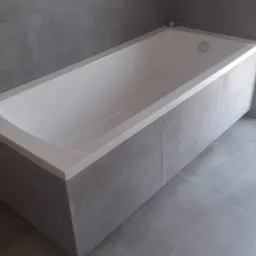 Nowa, biała wanna obudowana szarymi płytkami ceramicznymi, dopasowanymi do płytek na ścianie i podłodze. Widoczne niezabezpieczone przewody w ścianie.