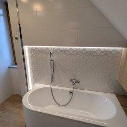 Daniel Falkowski - Nowoczesna łazienka na poddaszu z białą wanną, mozaiką 3D i subtelnym oświetleniem LED. Uwagę przykuwają drewnopodobne płytki podłogowe i okno w skosie.